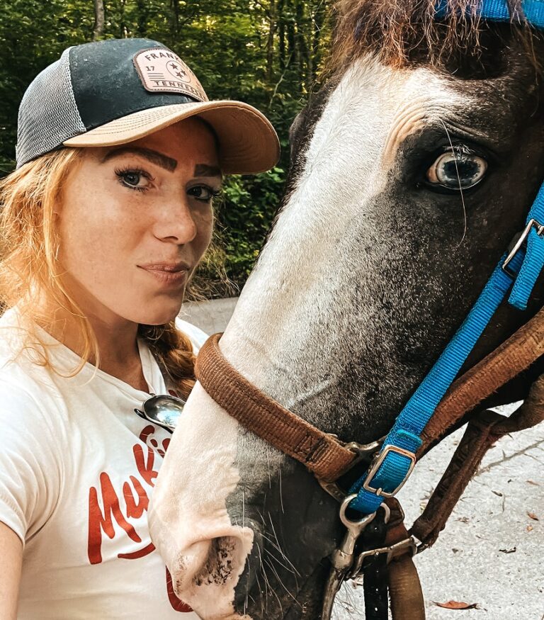 A woman wearing a cap and white t-shirt takes a close-up selfie with a horse wearing a blue halter outdoors.
