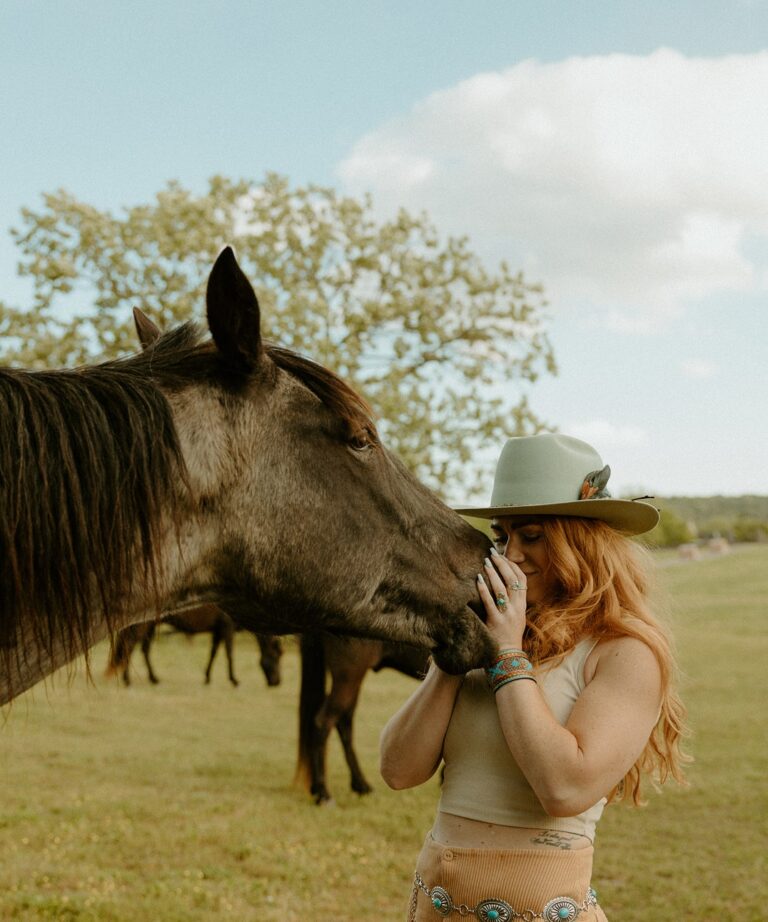 A woman in a green hat interacts closely with a horse in a grassy field, surrounded by trees and other horses in the background.