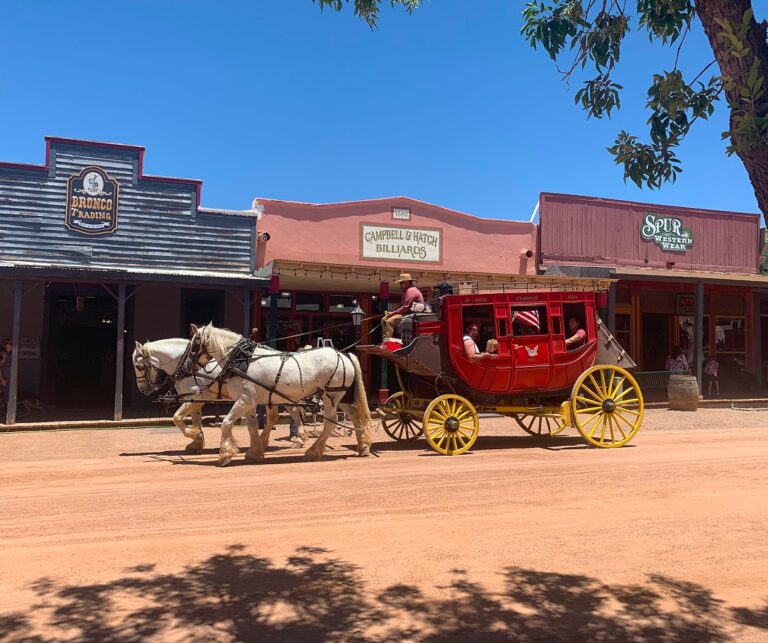 A horse-drawn stagecoach passes historic storefronts under a clear blue sky in a vibrant western town.