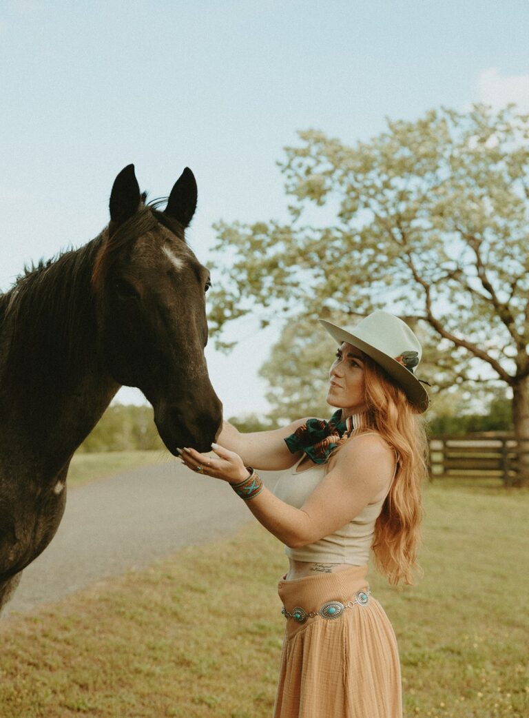 A woman in a cowboy hat gently interacts with a black horse, set against a serene rural landscape with trees and a gravel road.